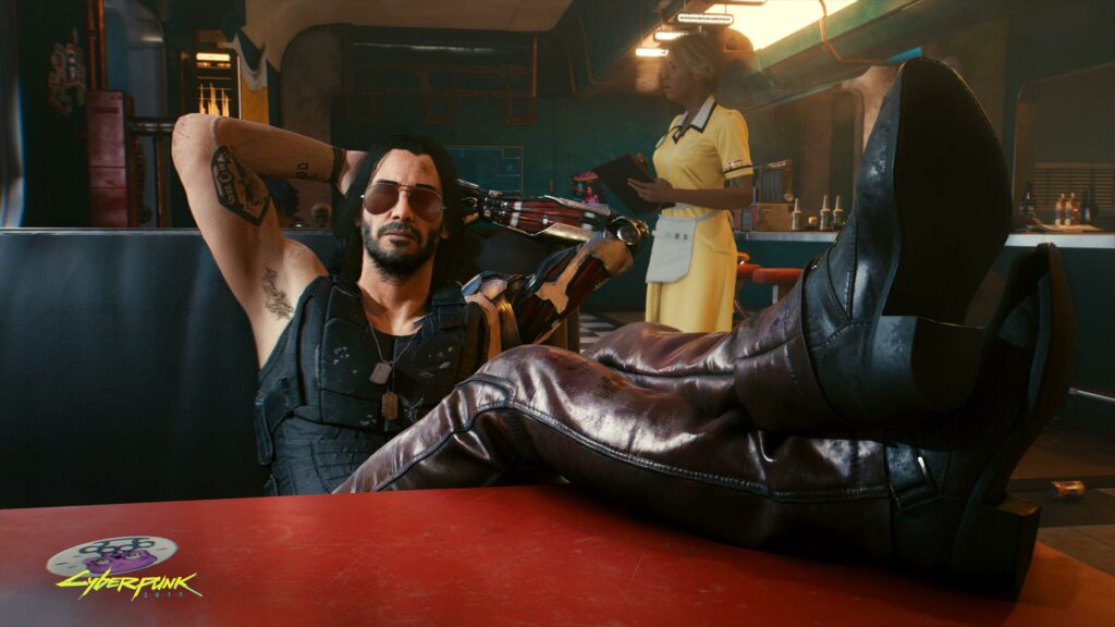 A screenshot from Cyberpunk 2077, a video game. The image depicts one of the main character's, Johnny Silverhand (played by and modeled after Keanu Reeves). The character is wearing leather pants, a bulletproof vest and sunglasses. He has his feet kicked up on a table in a diner.