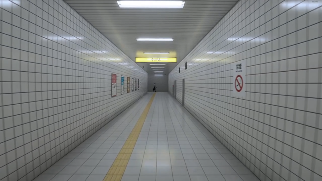 A screenshot from The Exit 8, a video game. The image depicts a liminal space. The space is a hallway made of white tiles. There are posters on the walls and a man wearing business casual attire at the end of the hallway.